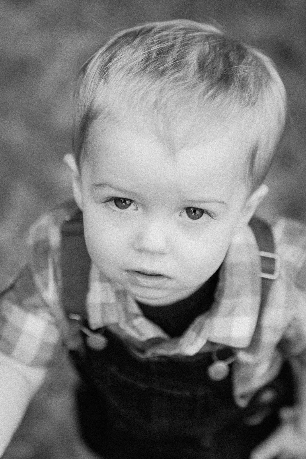 Black and white portrait of a young toddler boy in plaid shirt and overalls looking up at camera.