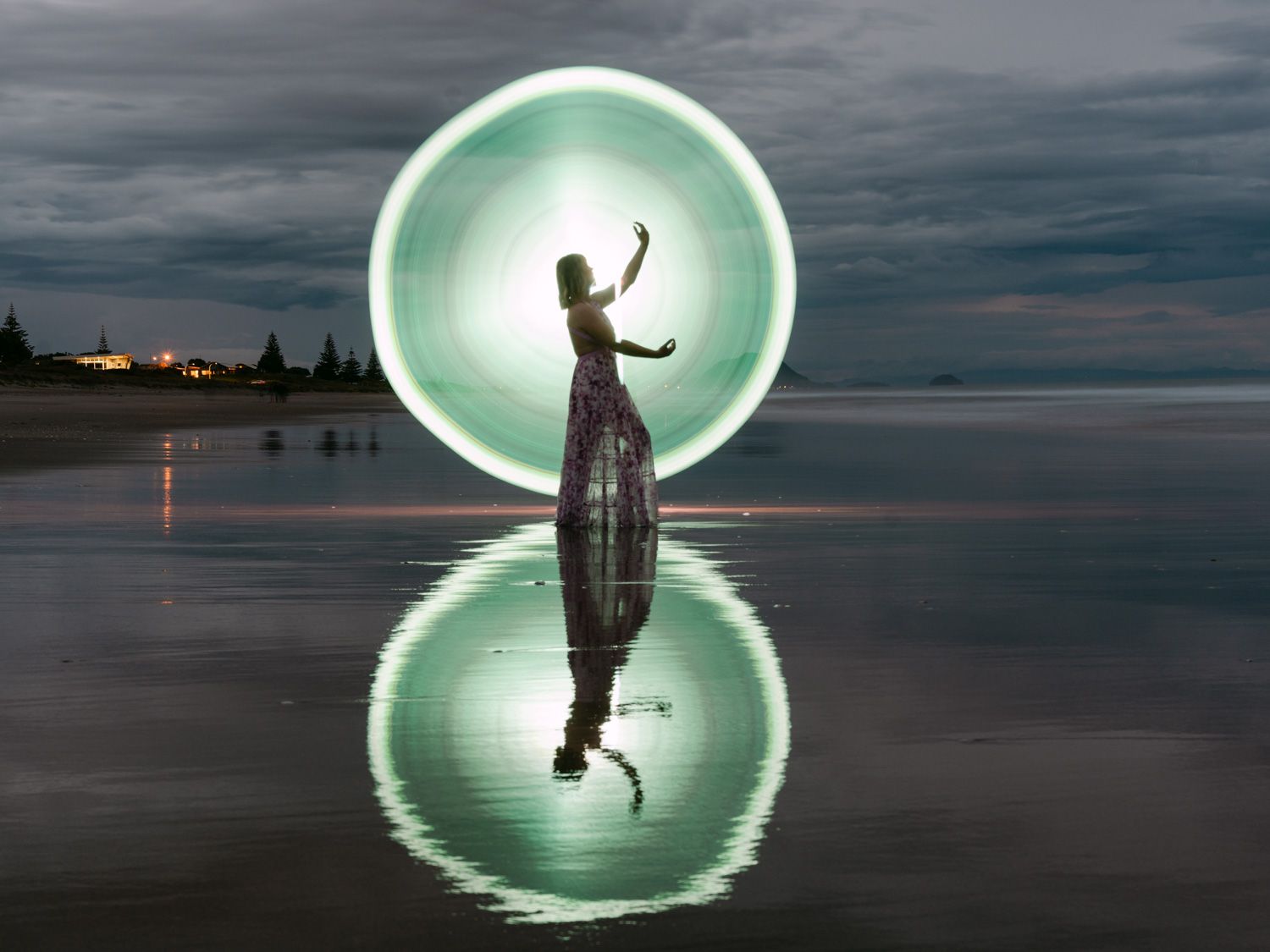 Woman standing on reflective beach with glowing green light circle behind her at dusk.