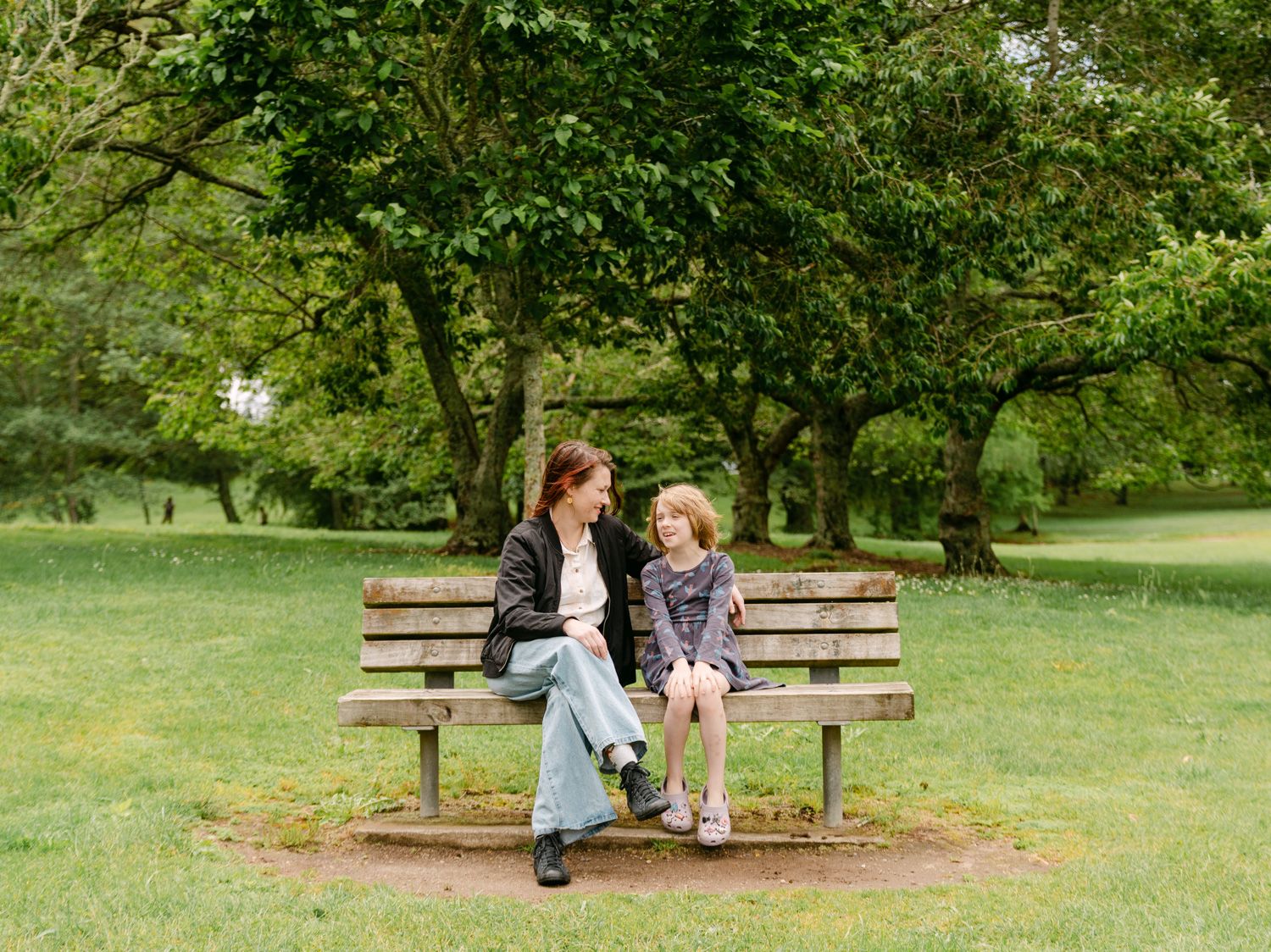 A woman and young girl sit together on a wooden park bench surrounded by lush green trees and grass.