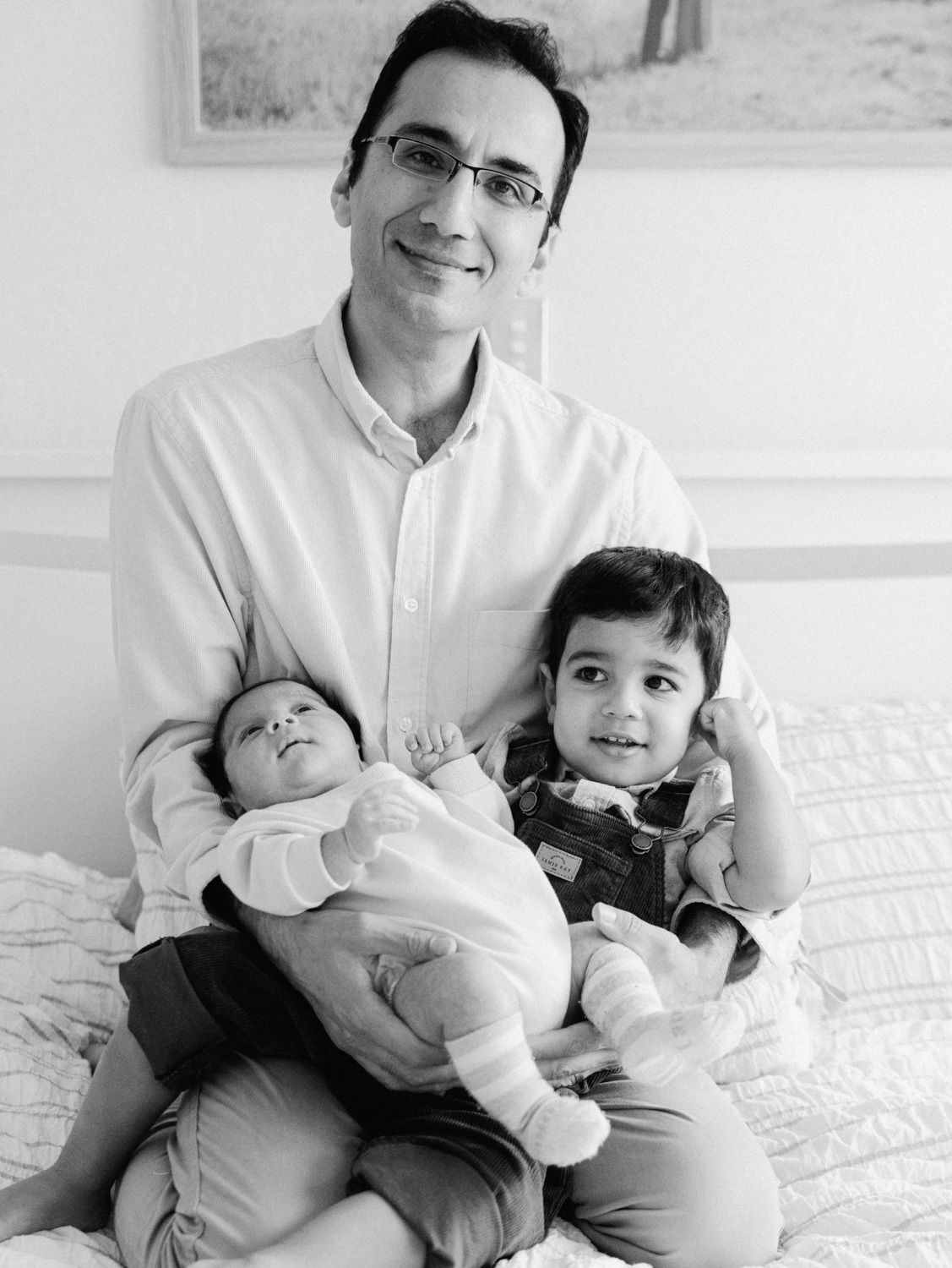 Black and white portrait of father holding newborn and toddler son while sitting on a bed.