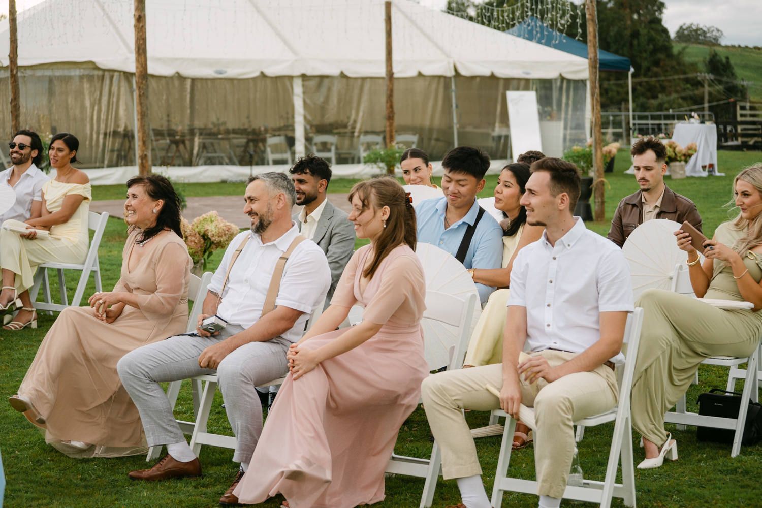 Wedding guests seated on white chairs outdoors, smiling and laughing during a ceremony on a green lawn.