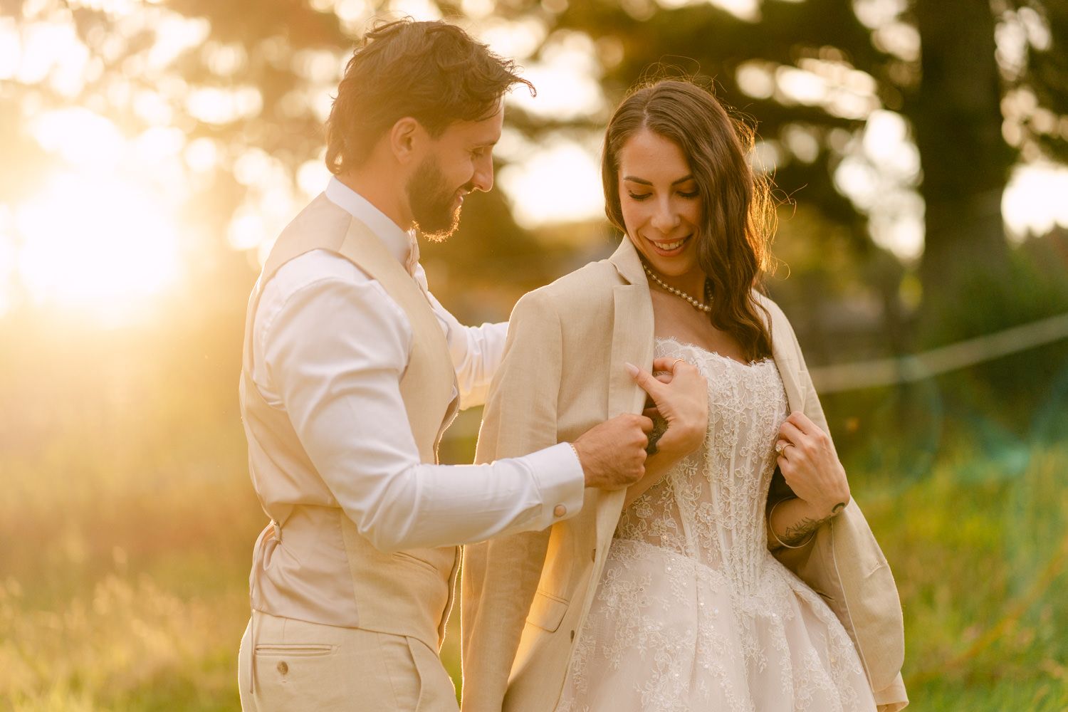 Groom placing jacket on bride in golden sunset light outdoors.