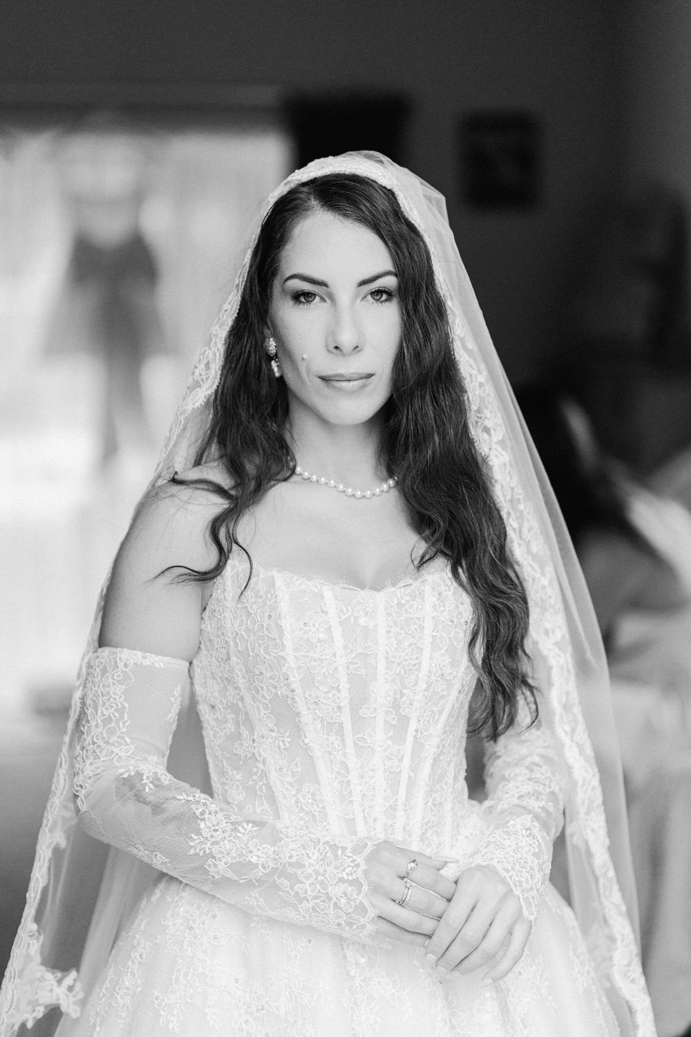 Black and white portrait of a beautiful bride in lace wedding gown and veil, wearing pearl necklace.