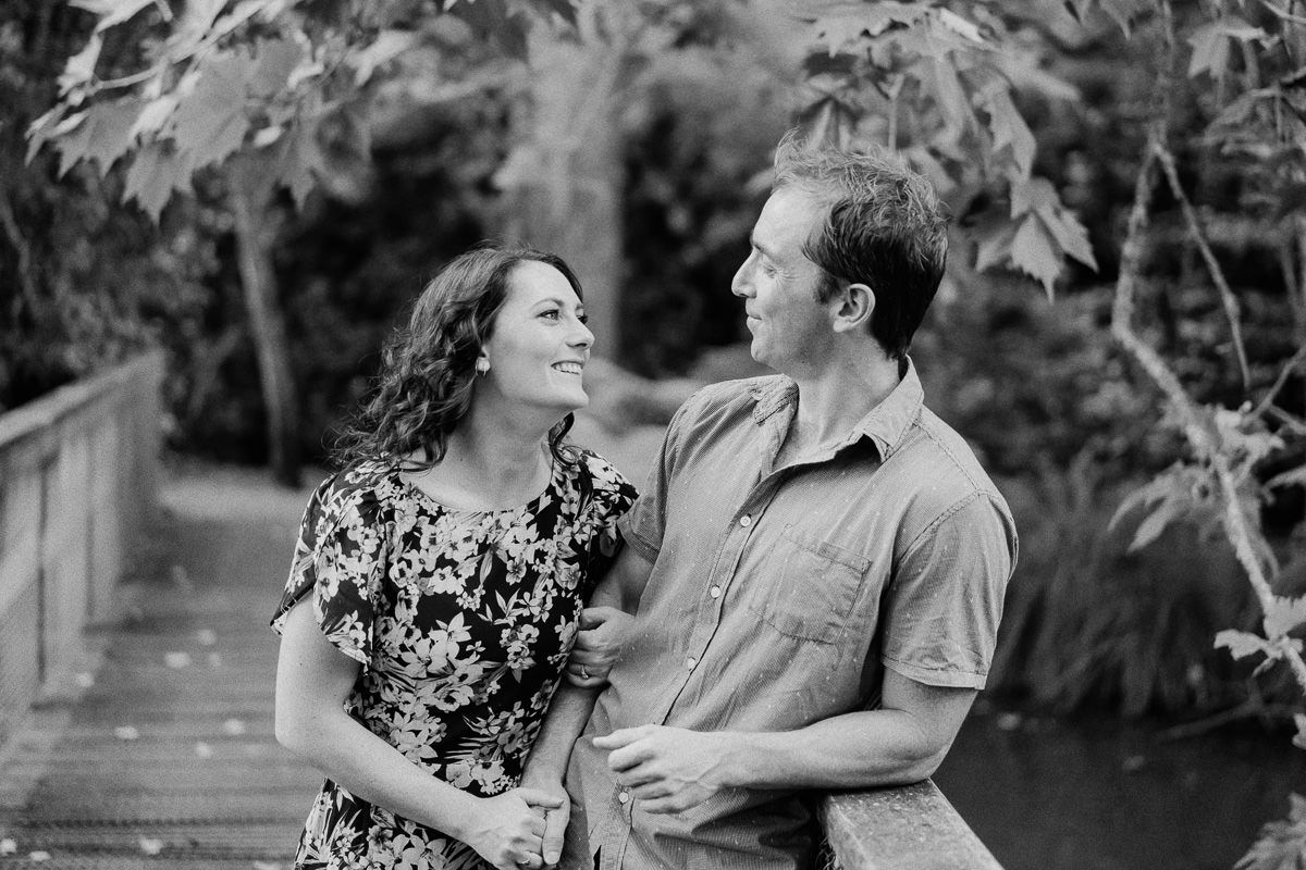 Black and white photo of a smiling couple gazing at each other outdoors near a wooden bridge.