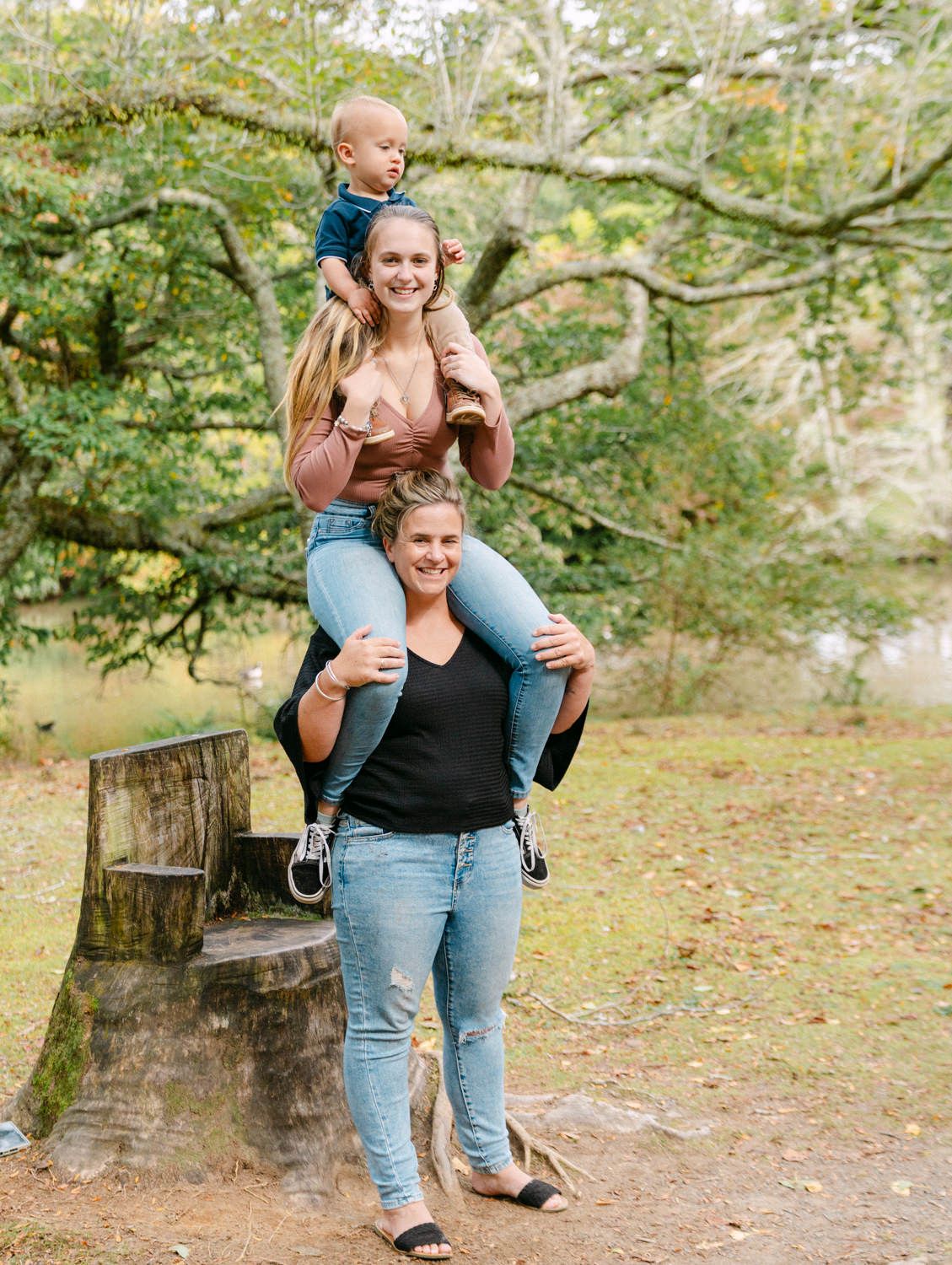 Two women and a toddler pose playfully outdoors, one carrying another on shoulders, in a lush green park setting.
