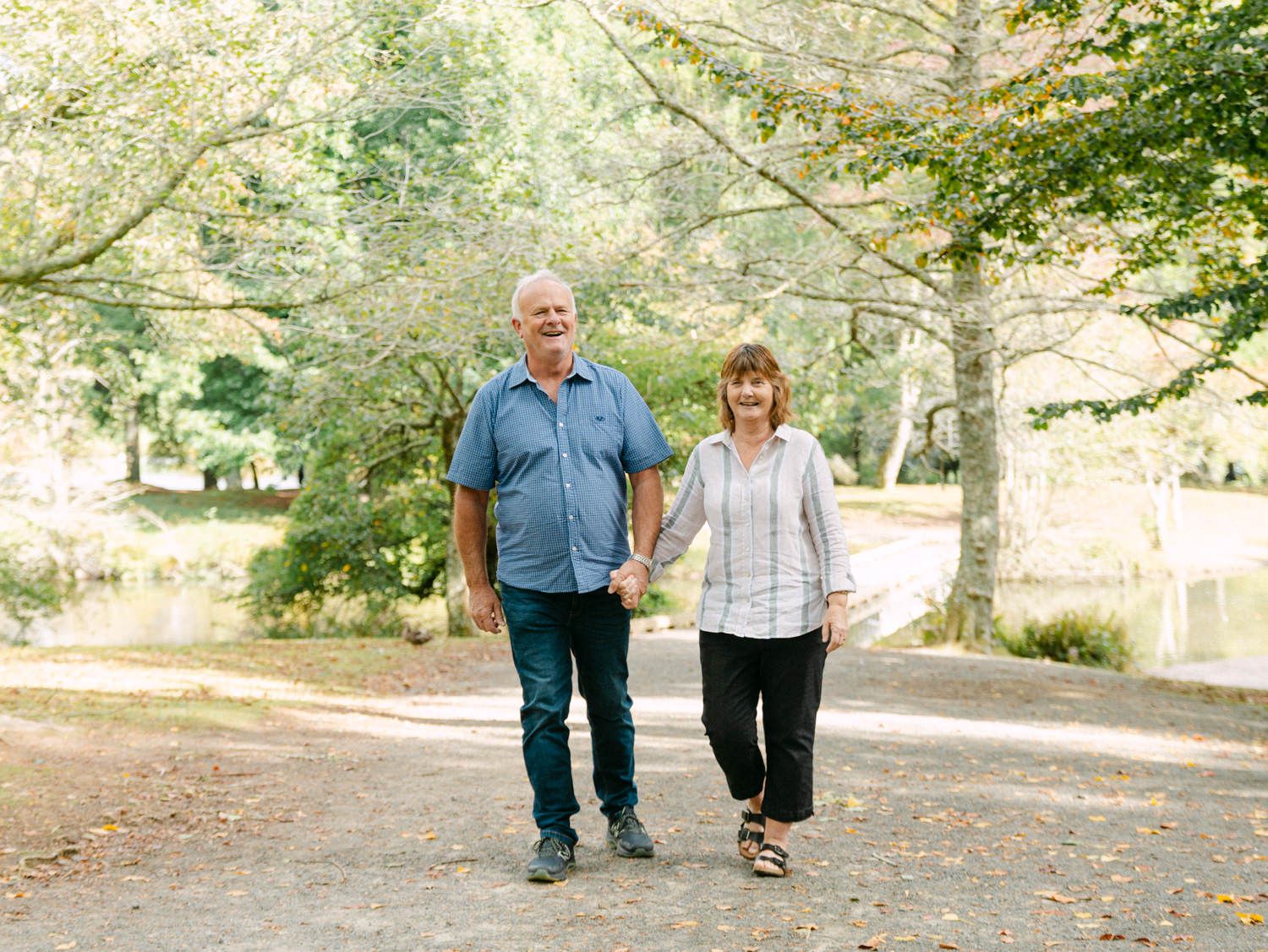 Elderly couple holding hands walking on a tree-lined park path on a sunny day.