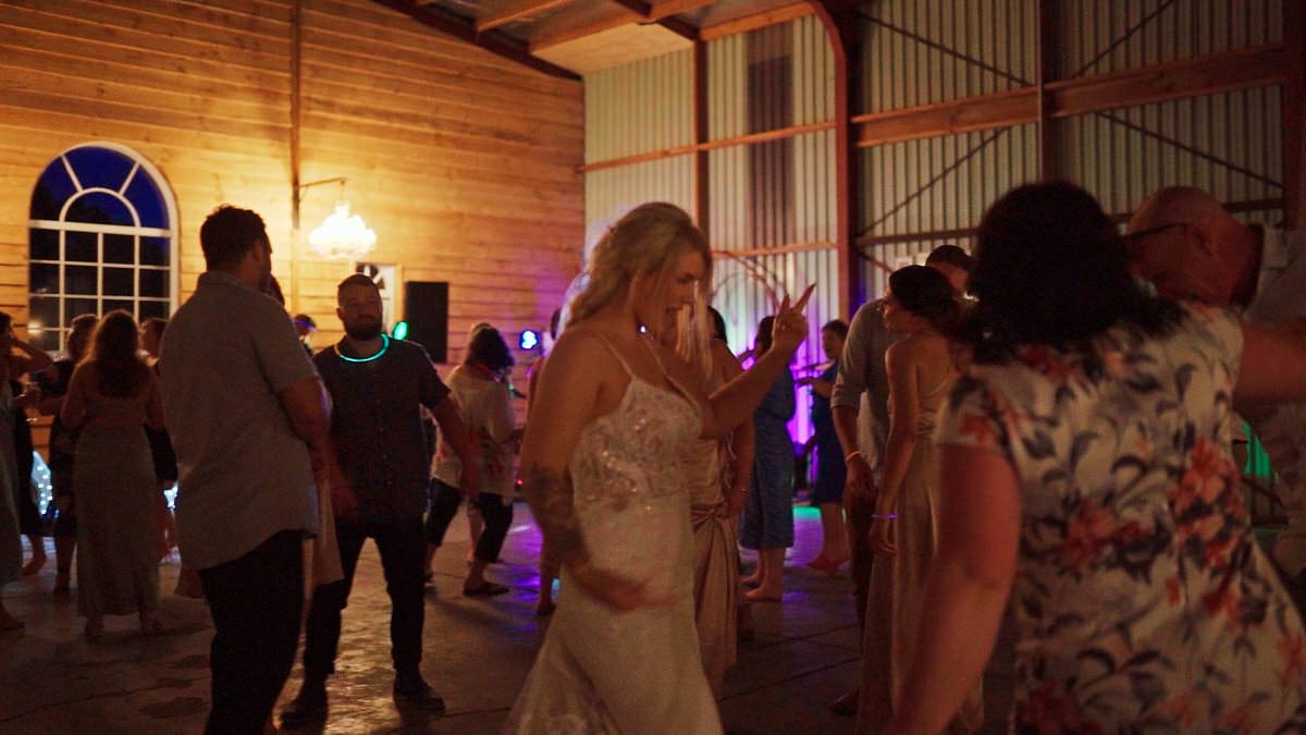 Bride in white wedding gown dancing with guests at a rustic barn wedding reception with colorful DJ lights.