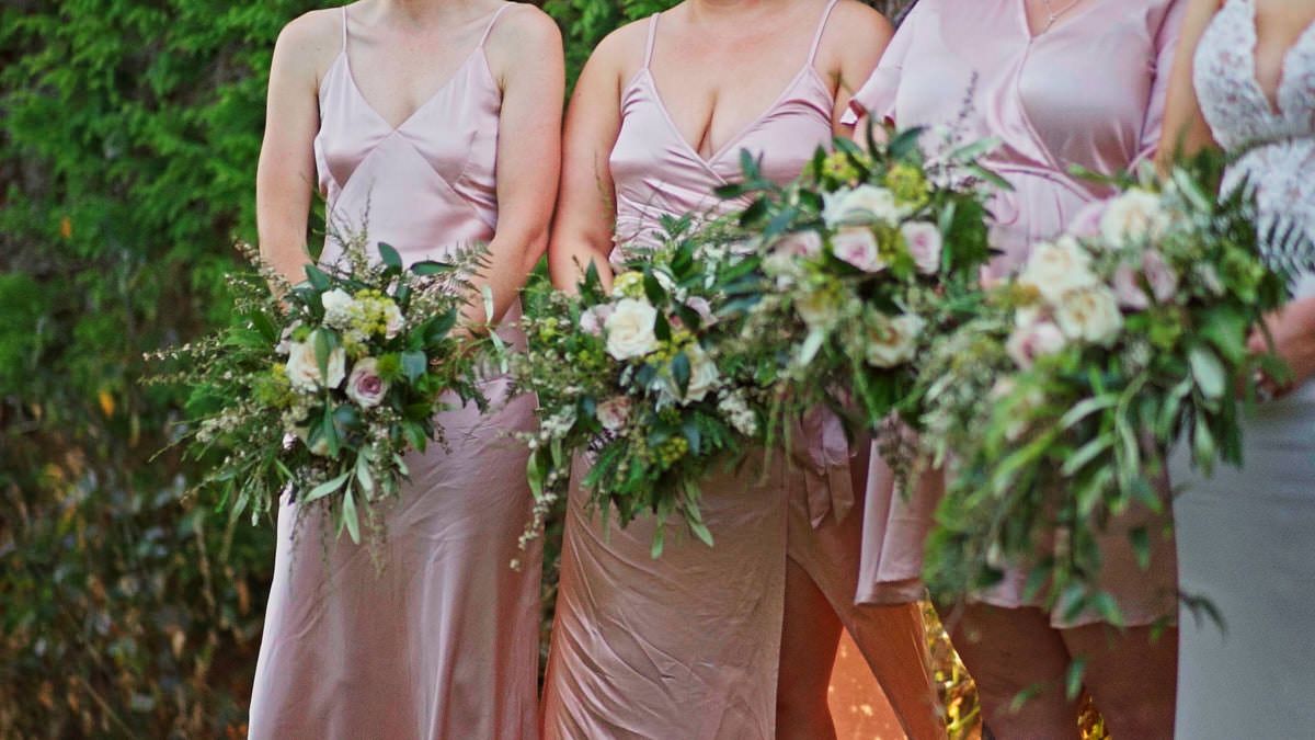 Bridesmaids in blush pink satin dresses holding lush white and green floral bouquets outdoors.