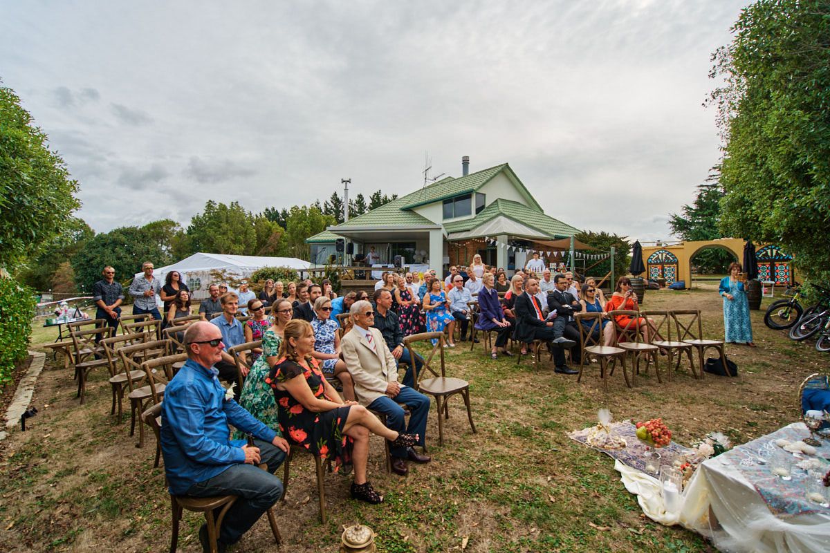 Outdoor gathering with guests seated on chairs in a garden near a blue house under a cloudy sky.