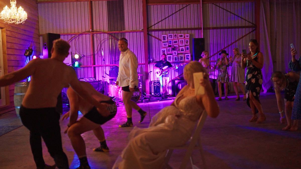 Wedding reception dance floor with bride in white dress, guests dancing, and live band performing on stage with purple lights.