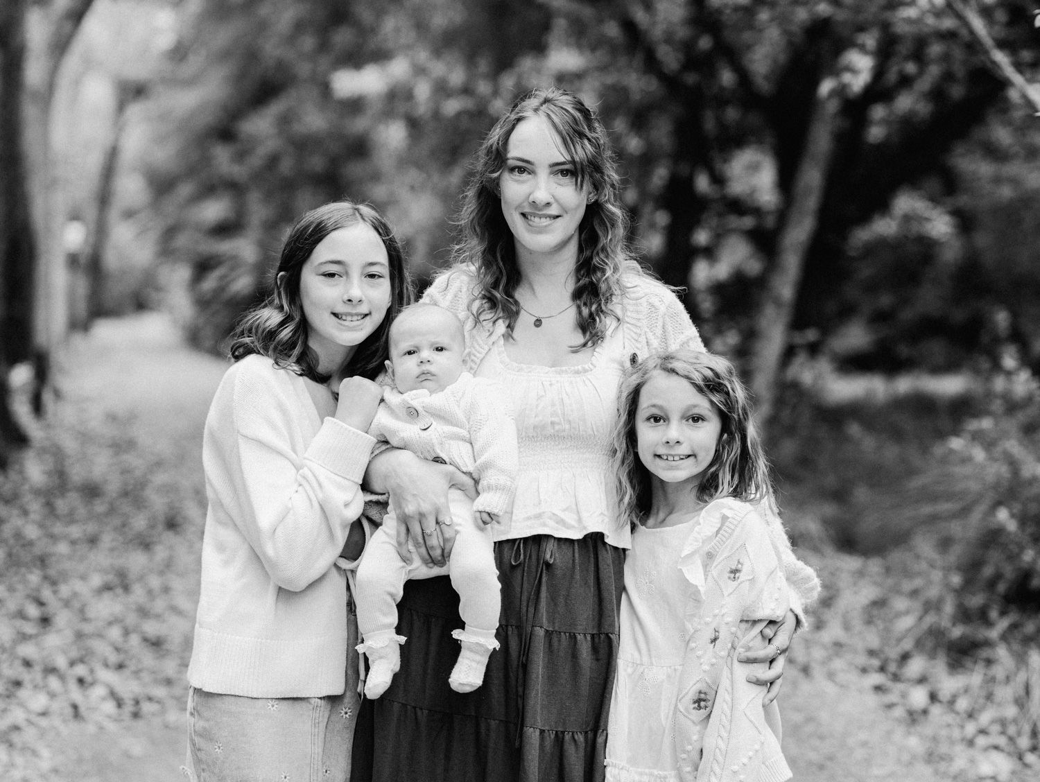 Black and white family portrait of a mother holding a baby with two daughters outdoors on a tree-lined path.