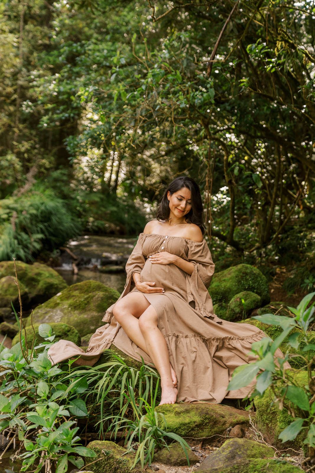 Pregnant woman in beige off-shoulder dress sitting on mossy rocks in lush green forest, cradling baby bump.