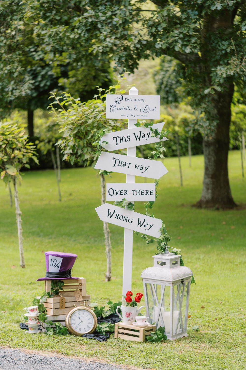 Alice in Wonderland wedding sign with directional arrows, Mad Hatter hat, clock, and lantern decor on green lawn.
