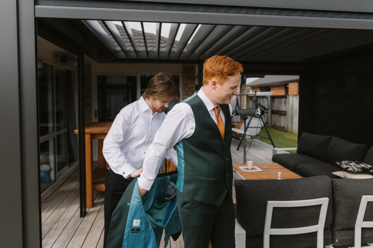 Two groomsmen in green suits and orange ties prepare for a wedding on an outdoor deck.