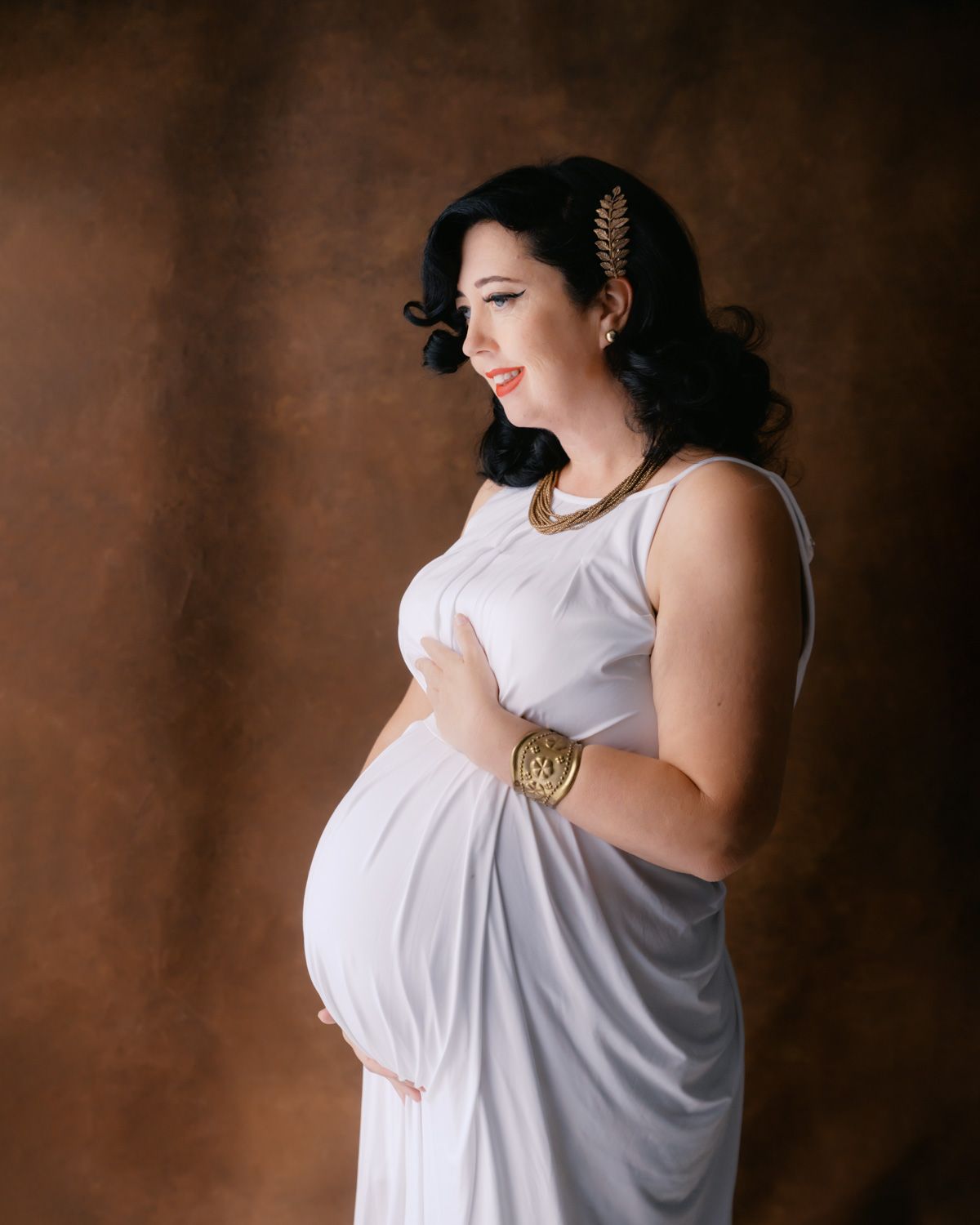 Pregnant woman in white Grecian gown with gold accessories poses gracefully in studio maternity portrait.