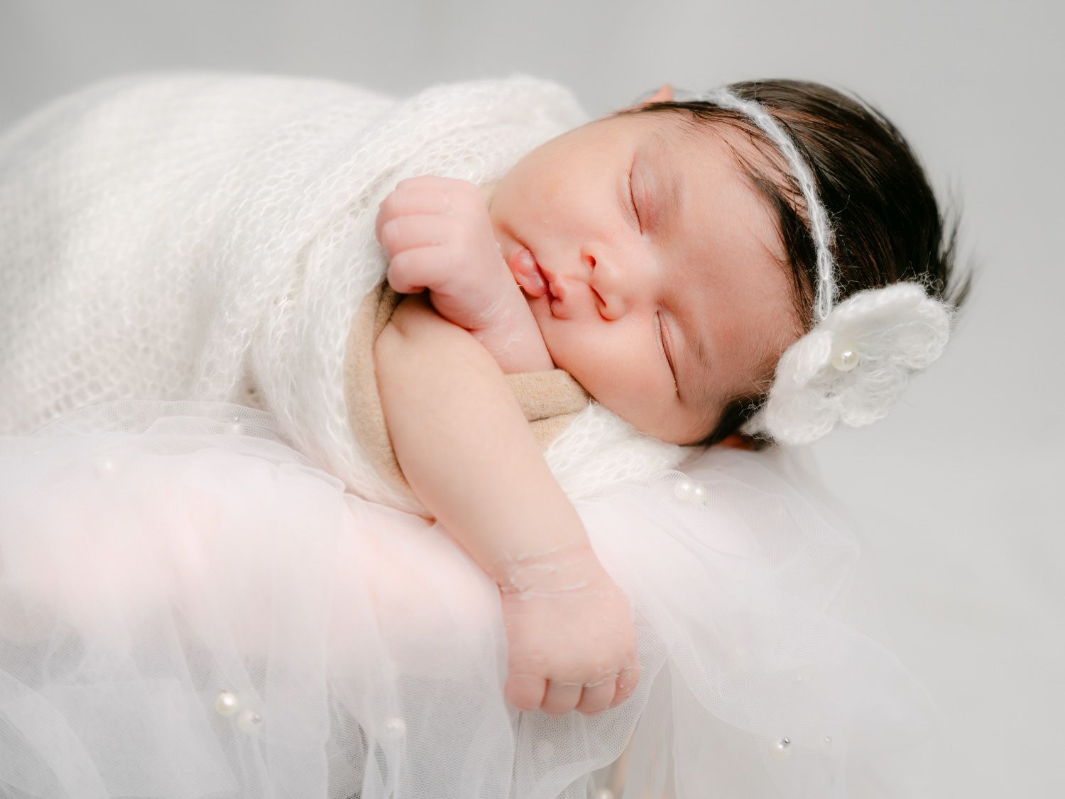 Peaceful newborn baby girl sleeping wrapped in white knit swaddle with floral headband on soft white background.