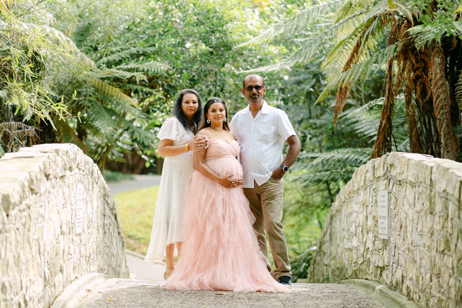 Pregnant woman in pink tulle gown poses with family on stone bridge surrounded by lush green tropical foliage.