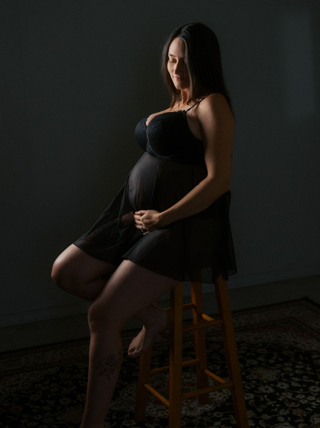 Pregnant woman in black dress sitting on wooden stool, cradling baby bump in moody studio lighting.