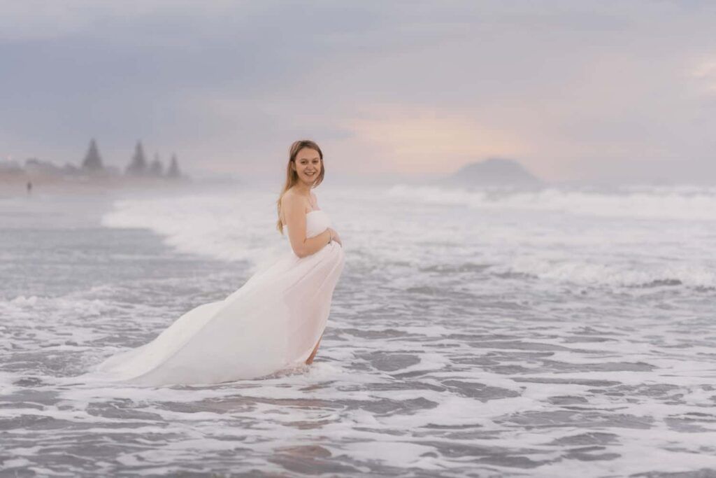 Bride in white gown standing in ocean waves at sunset on a misty beach.