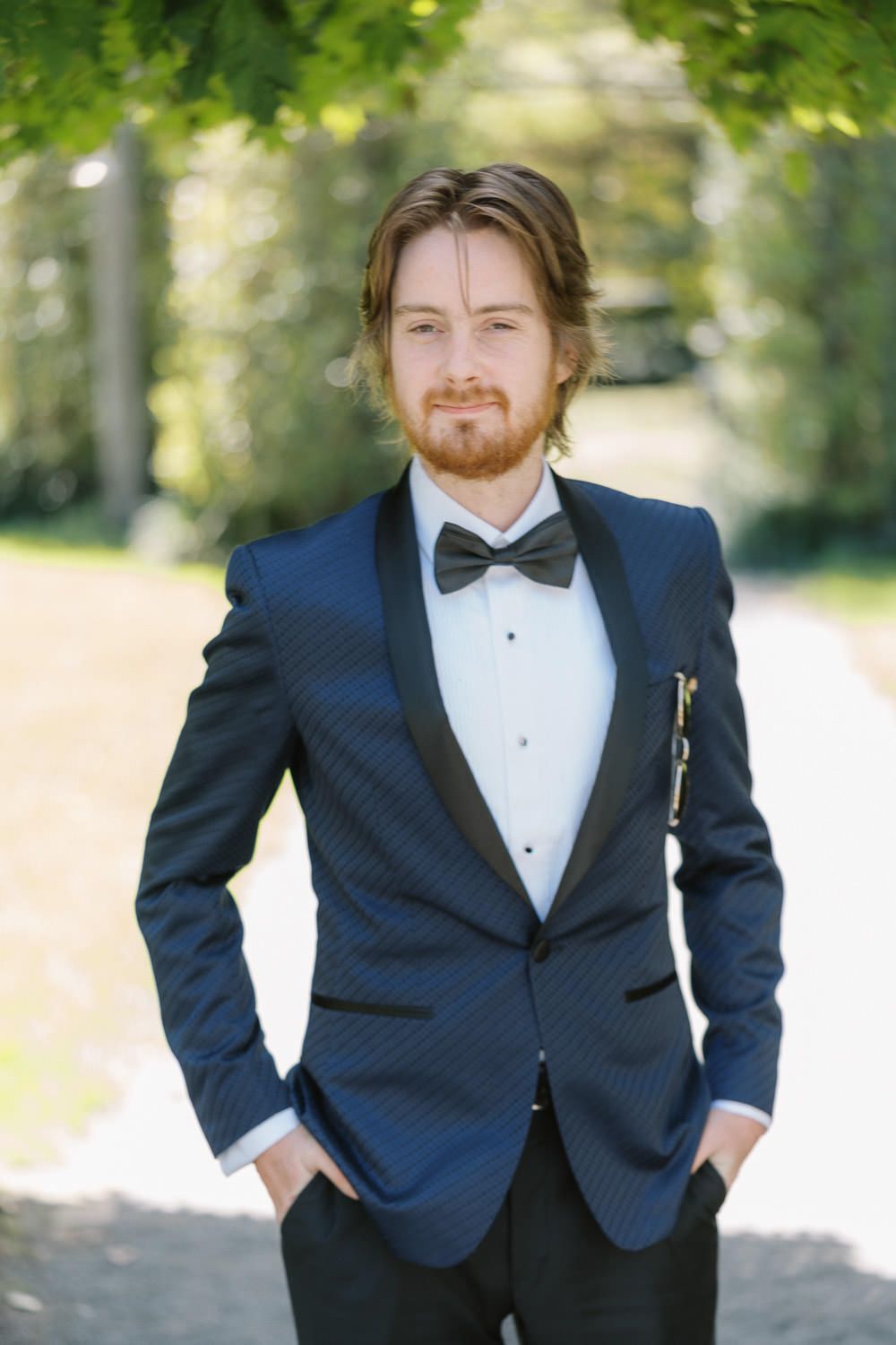 Elegant man in navy tuxedo jacket with black bow tie and pocket square, posing outdoors in a lush green garden.