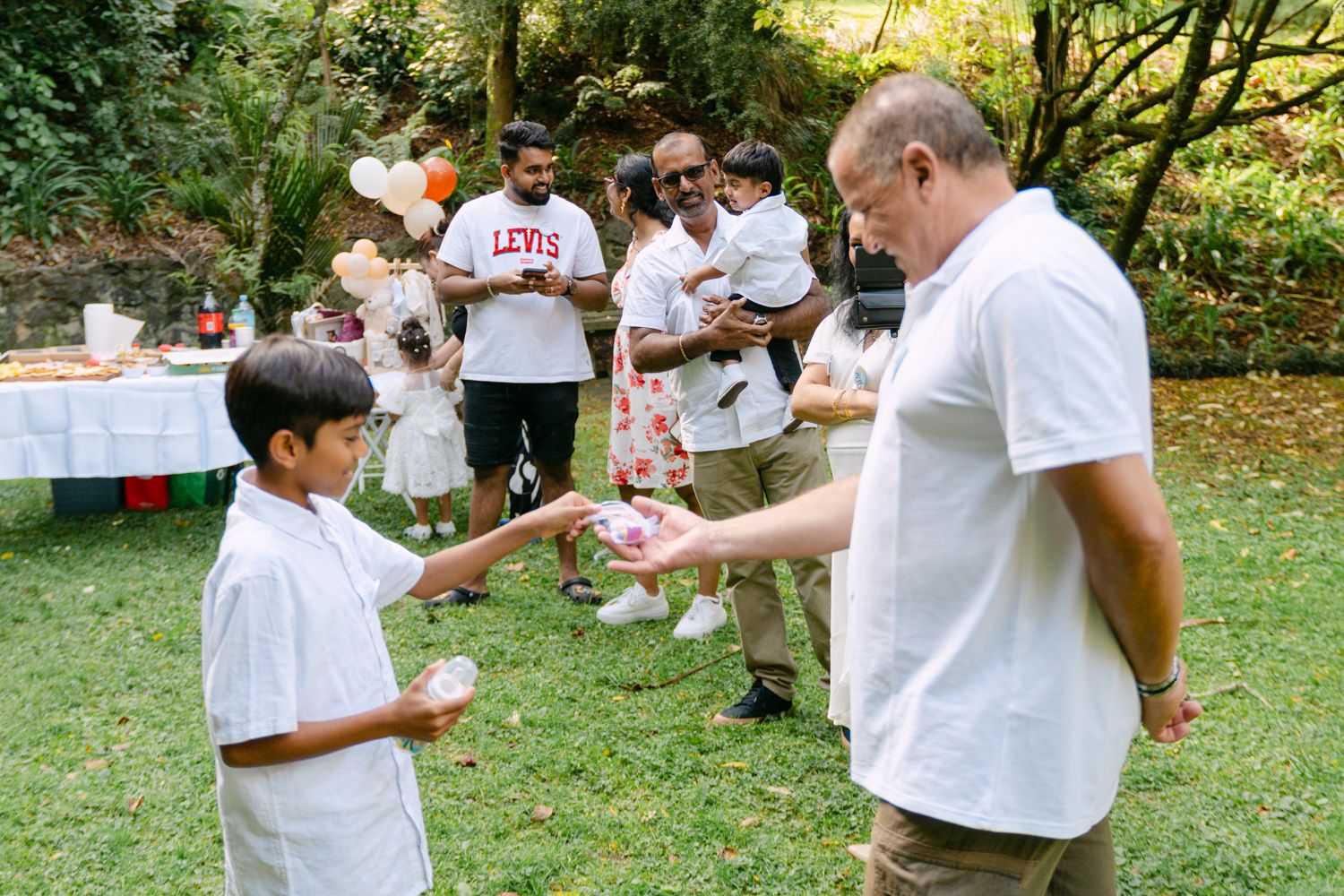 Man in white shirt handing gift to young boy at outdoor garden party with family gathered in background.