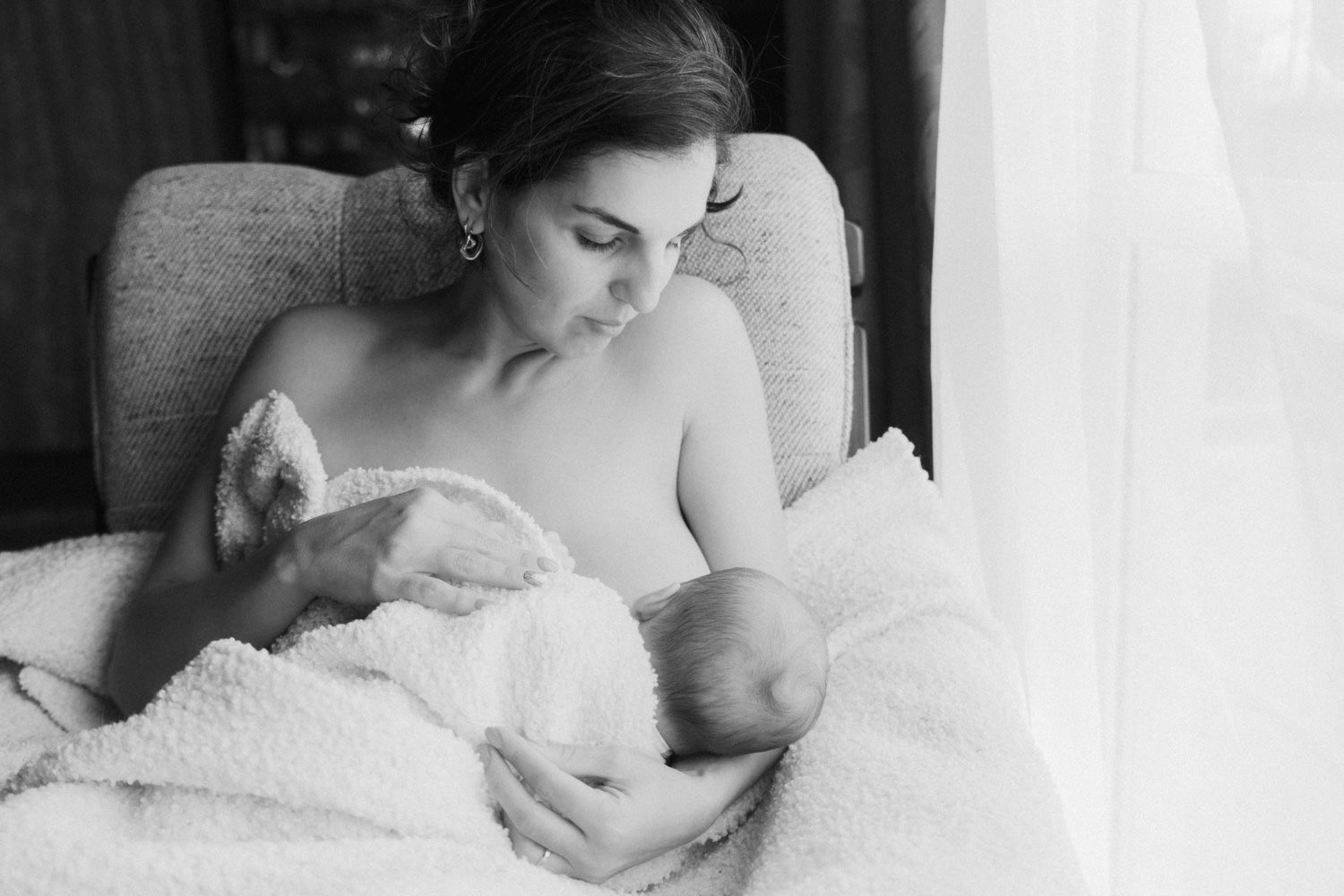 Black and white photo of a mother breastfeeding her newborn baby wrapped in a soft blanket near a window.
