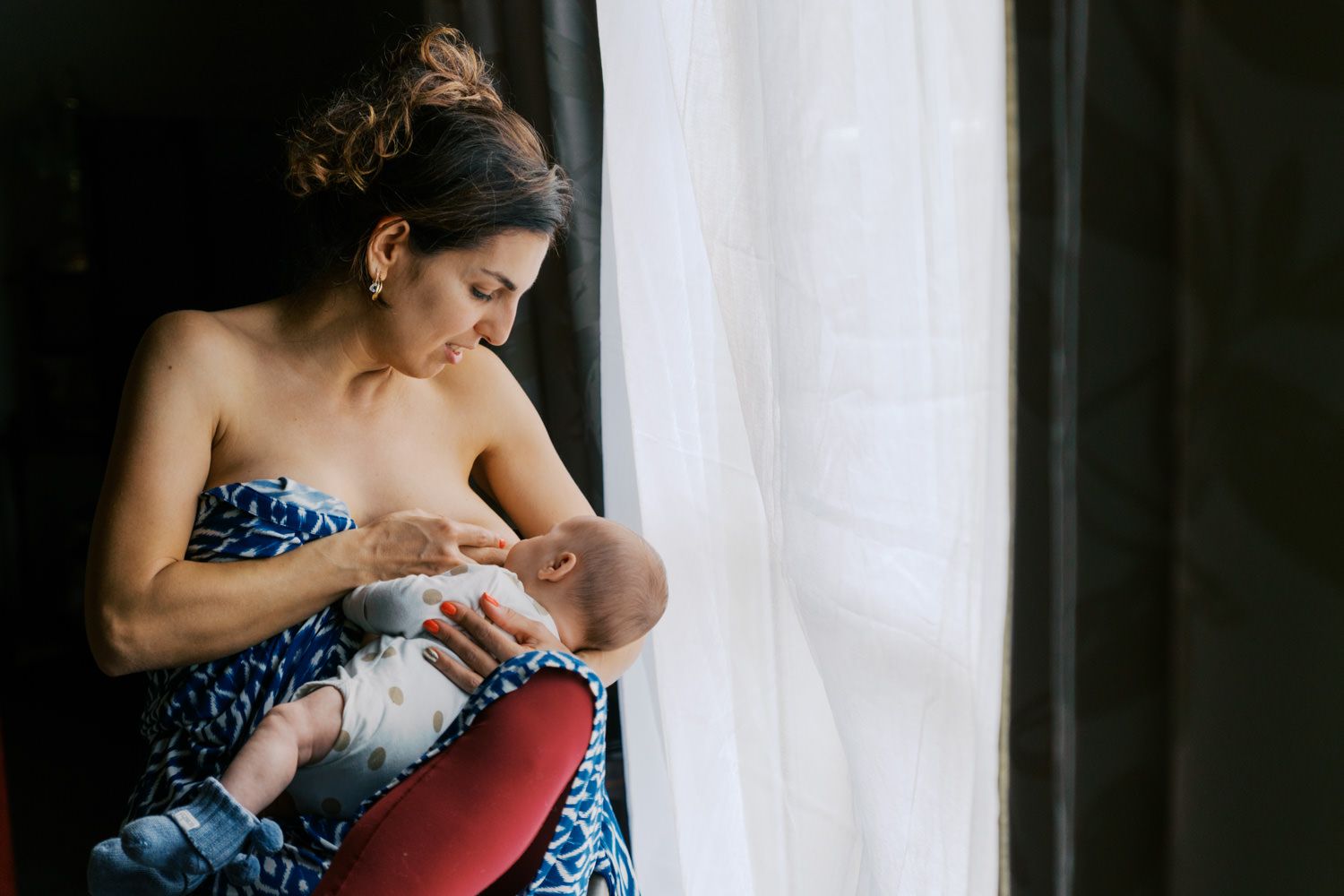 Mother breastfeeding newborn baby by a sunlit window, wearing a blue floral wrap dress in a serene indoor setting.