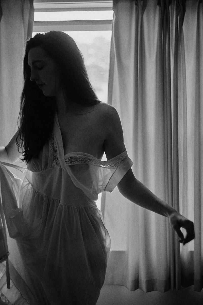 Black and white photo of a woman in a sheer white dress standing by a curtained window.