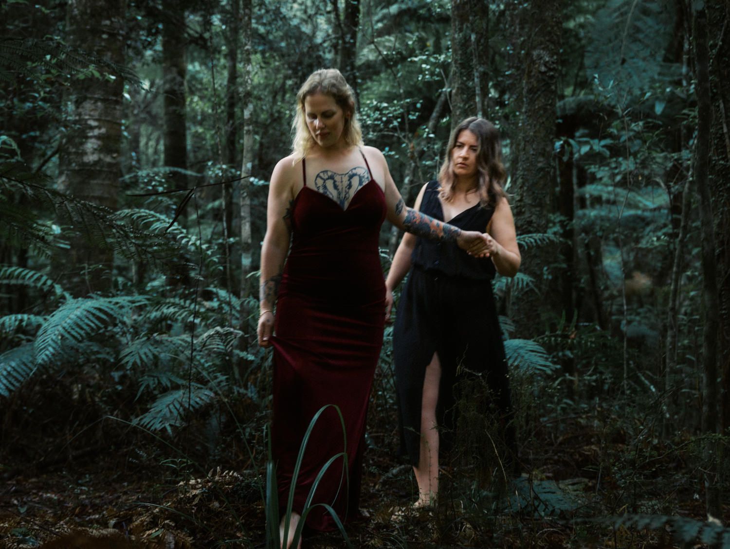 Two women in elegant dresses walk through a lush green forest in a moody, atmospheric outdoor photoshoot.