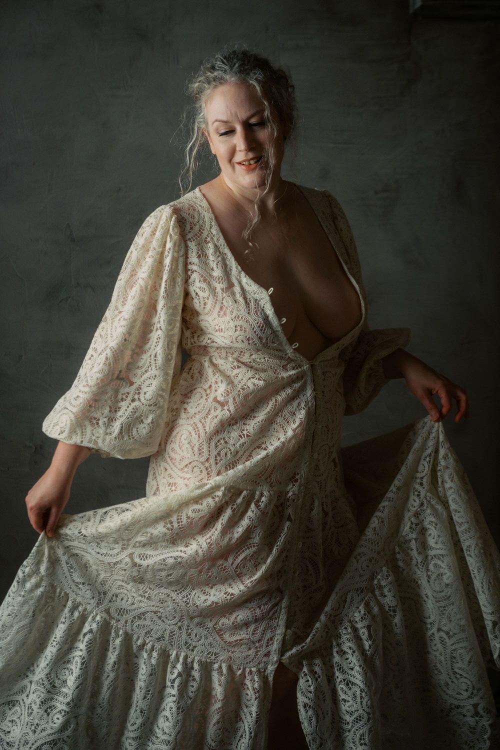 Woman in elegant cream lace gown smiling, holding skirt in moody studio setting.