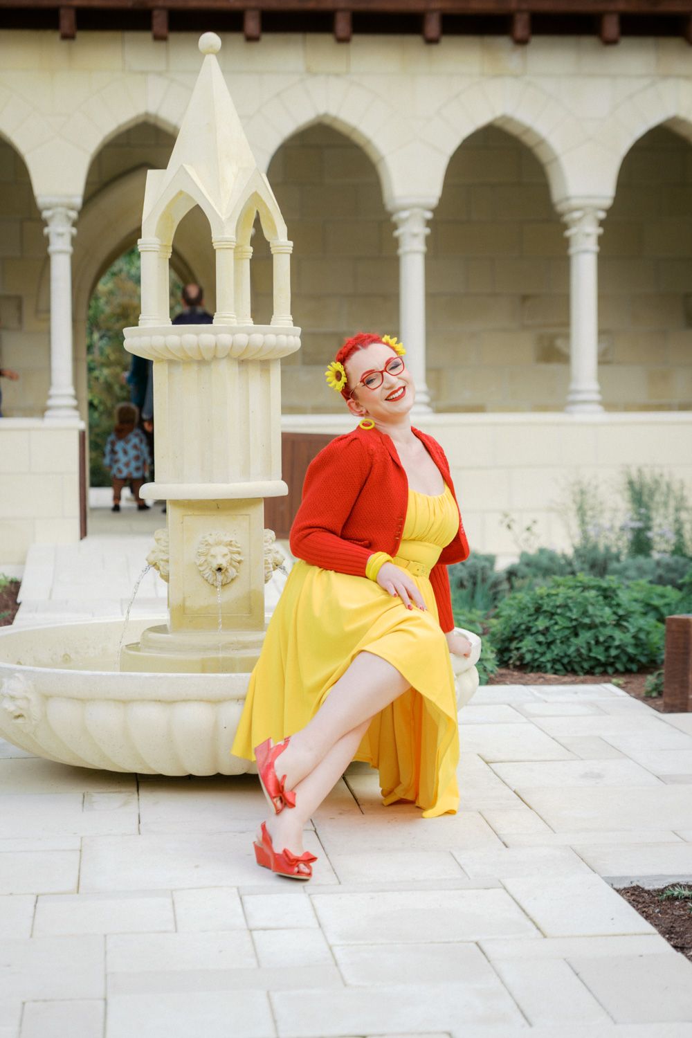 Woman in yellow dress and red cardigan posing by ornate fountain in elegant courtyard with arched colonnade.