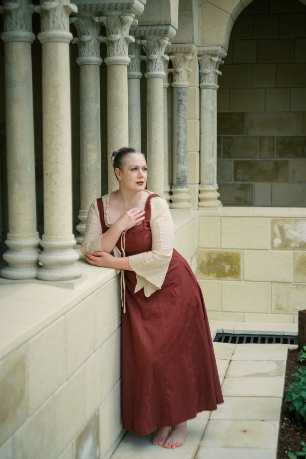 Woman in medieval burgundy dress leans against stone columns in historic courtyard, gazing thoughtfully into distance.