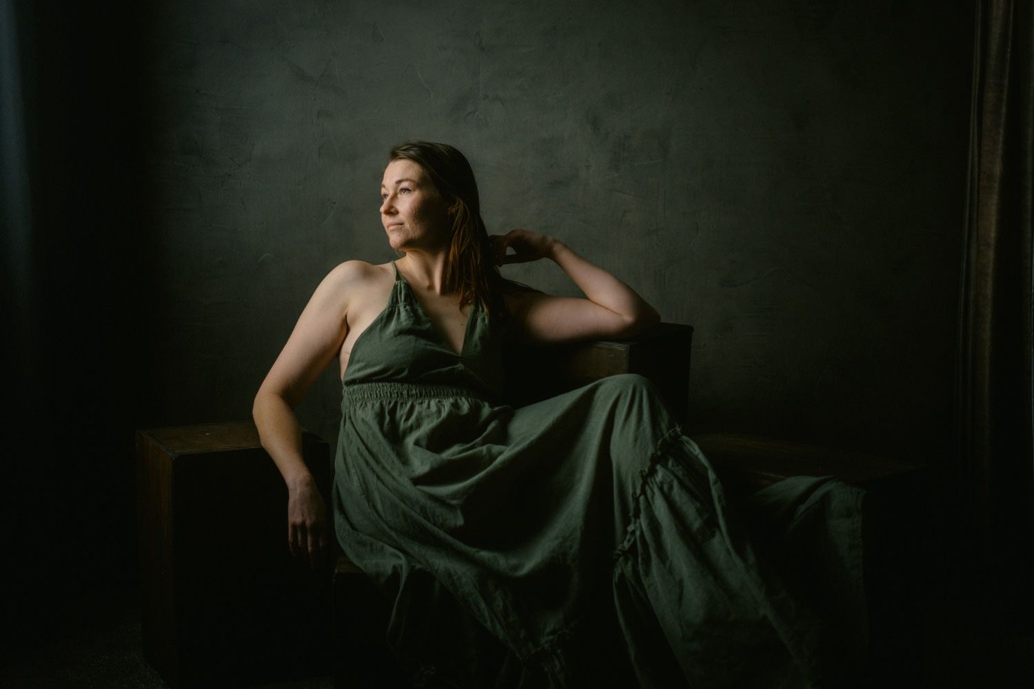 Woman in elegant green dress seated in moody studio lighting, looking away.