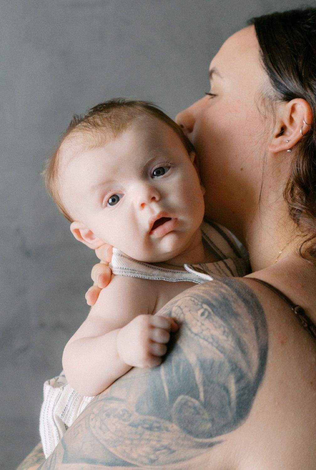 A tattooed parent tenderly holds a wide-eyed baby against a gray background.