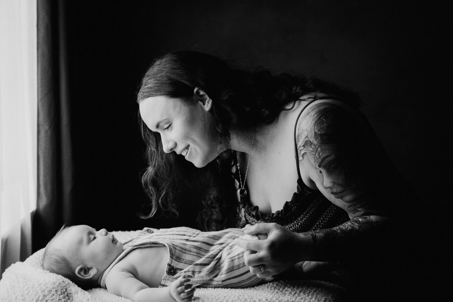 Black and white photo of tattooed mother smiling at her newborn baby lying on a blanket near a window.