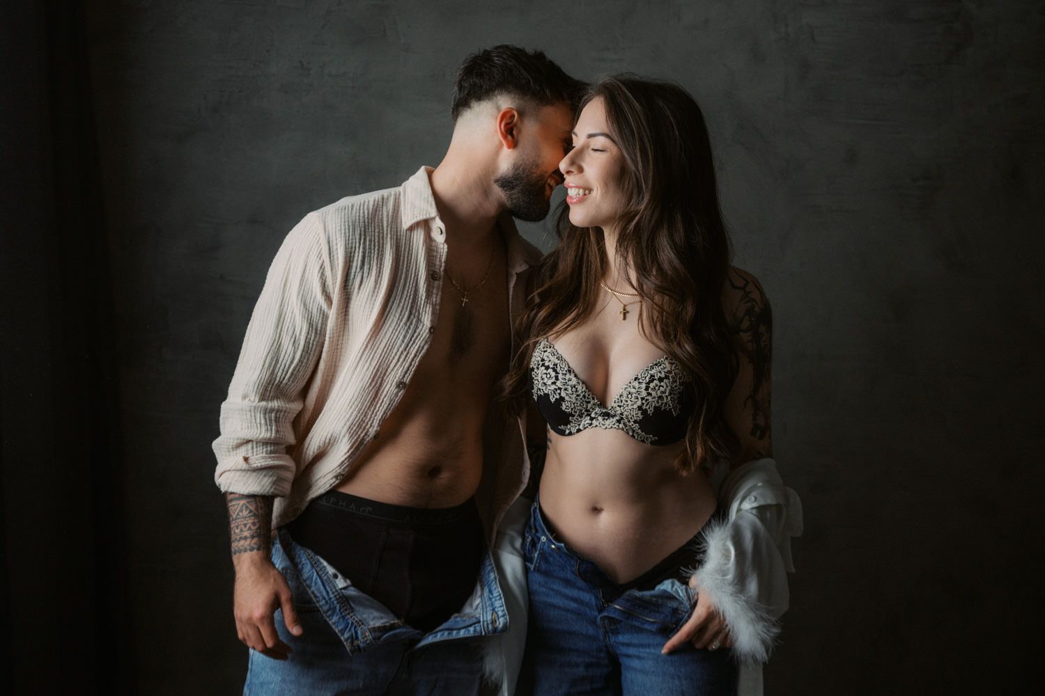 A romantic couple in casual denim and lingerie share an intimate moment against a dark studio backdrop.