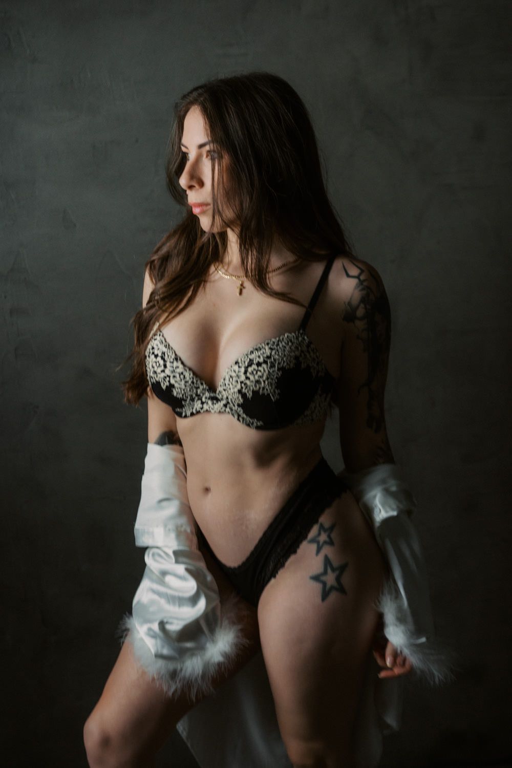 Woman in black lace lingerie and white robe with fur cuffs, featuring star tattoos, posing against dark studio backdrop.