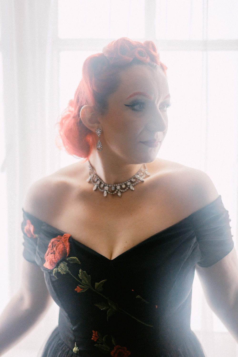 Woman in black off-shoulder floral gown with red updo hair, crystal necklace, posing elegantly near bright window.