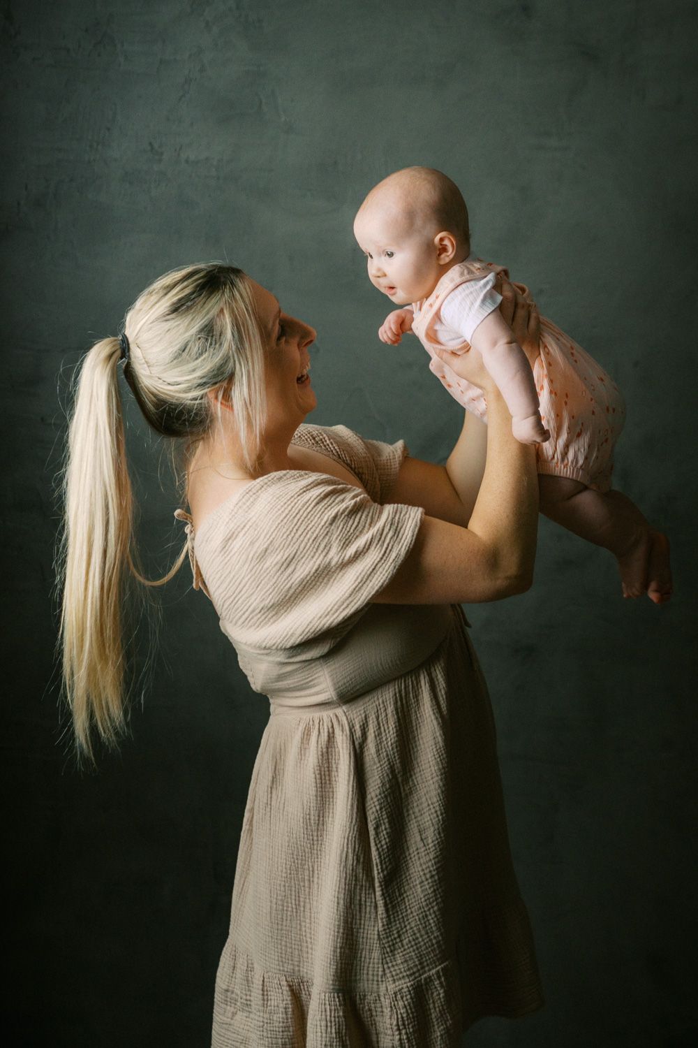 A blonde mother in a beige dress lifts her smiling baby in a pink outfit against a dark studio background.