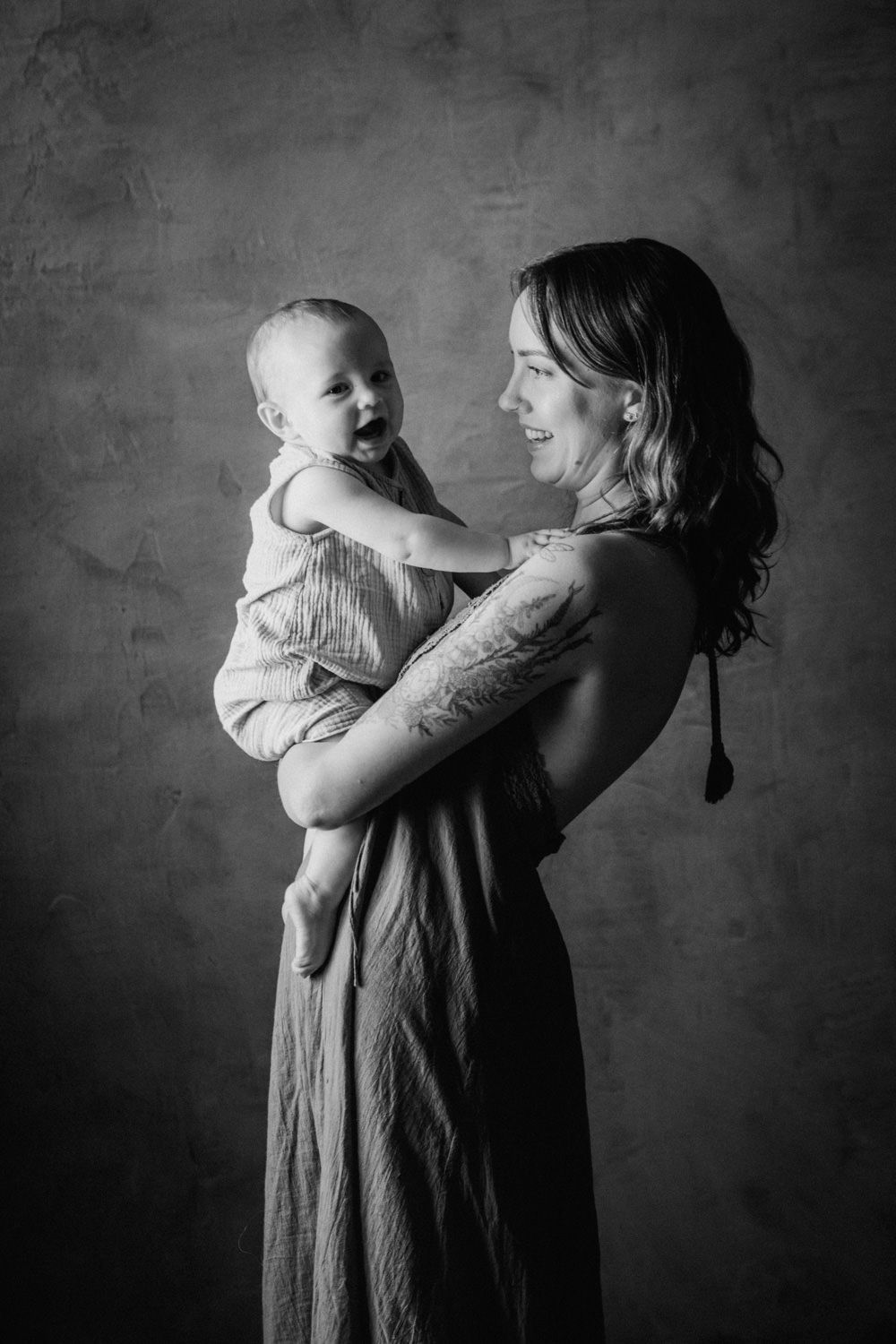 Black and white studio portrait of a smiling mother holding her laughing baby, showcasing a tender maternal bond.