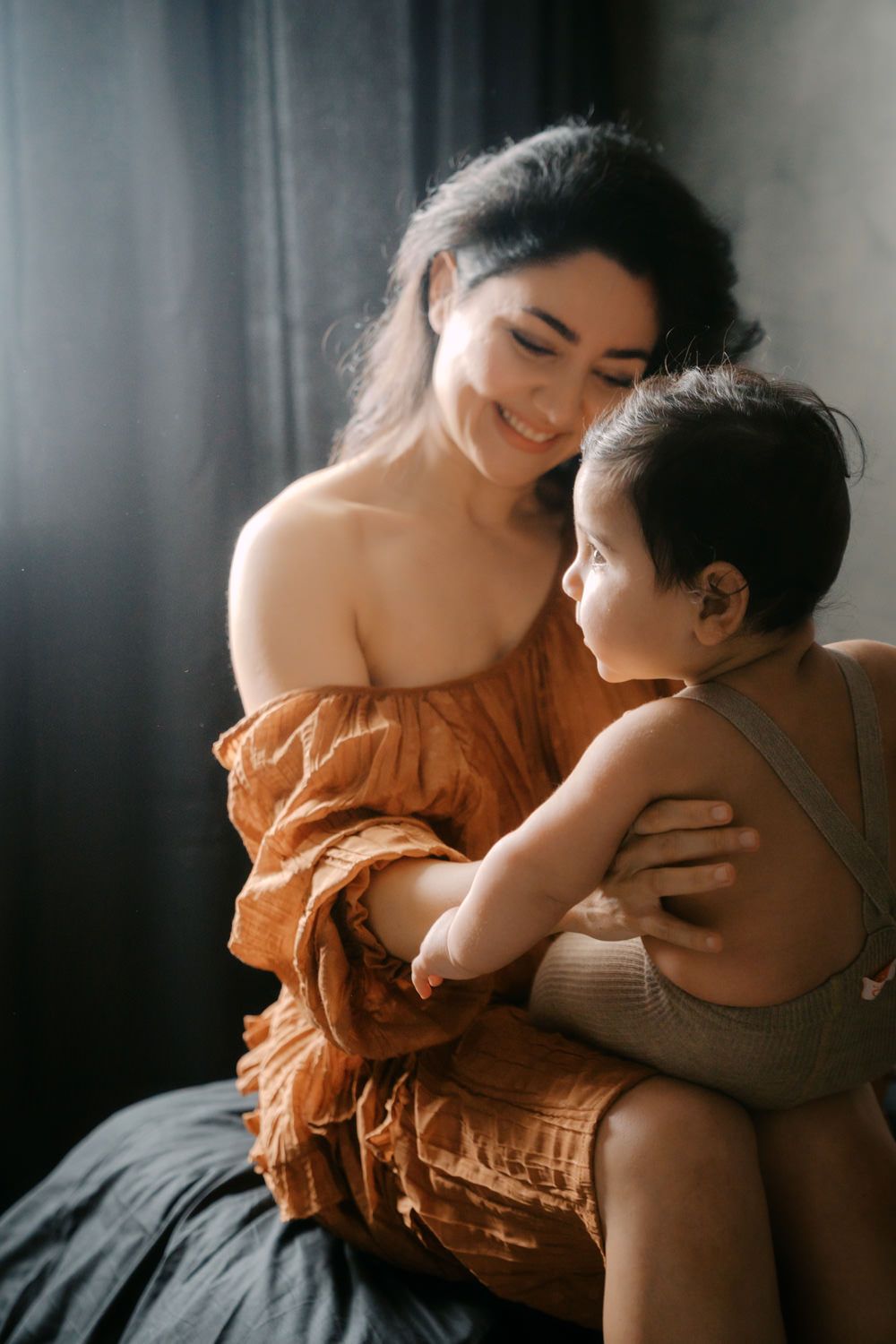 A smiling young mother in a rust-colored off-shoulder dress lovingly holds her toddler in a warmly lit room.