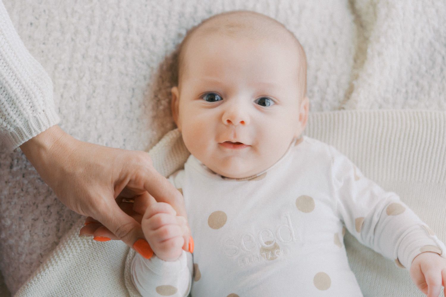 Adorable baby in polka dot onesie lying on white blanket, holding an adult's finger with blue eyes gazing up.
