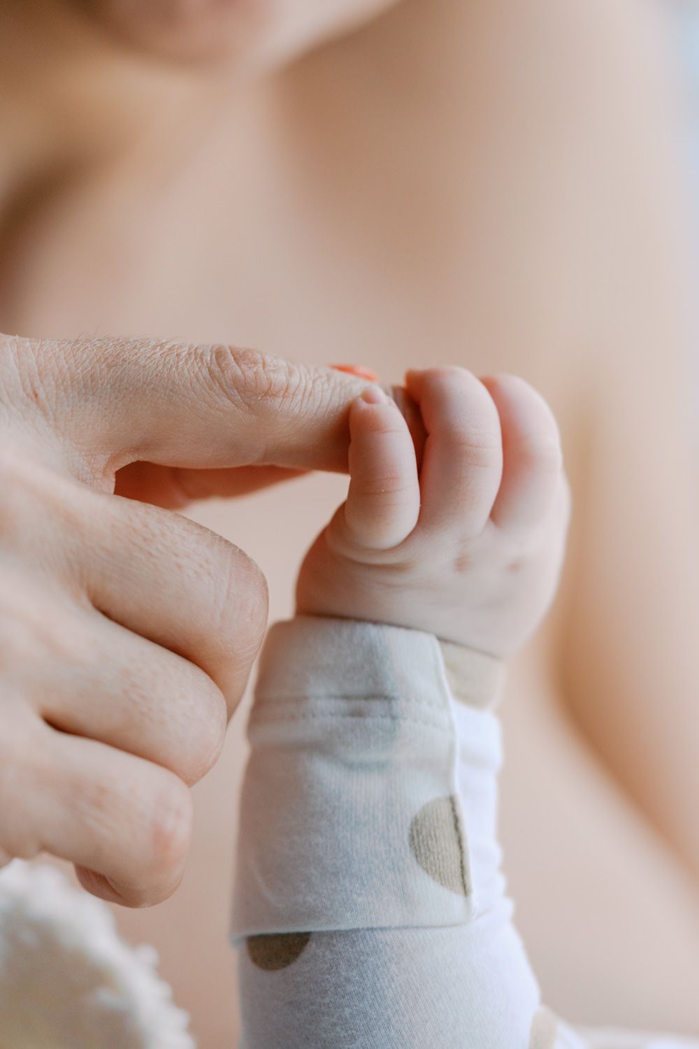 Close-up of a baby's tiny hand grasping an adult's finger, with infant wearing a white polka dot sleeve.