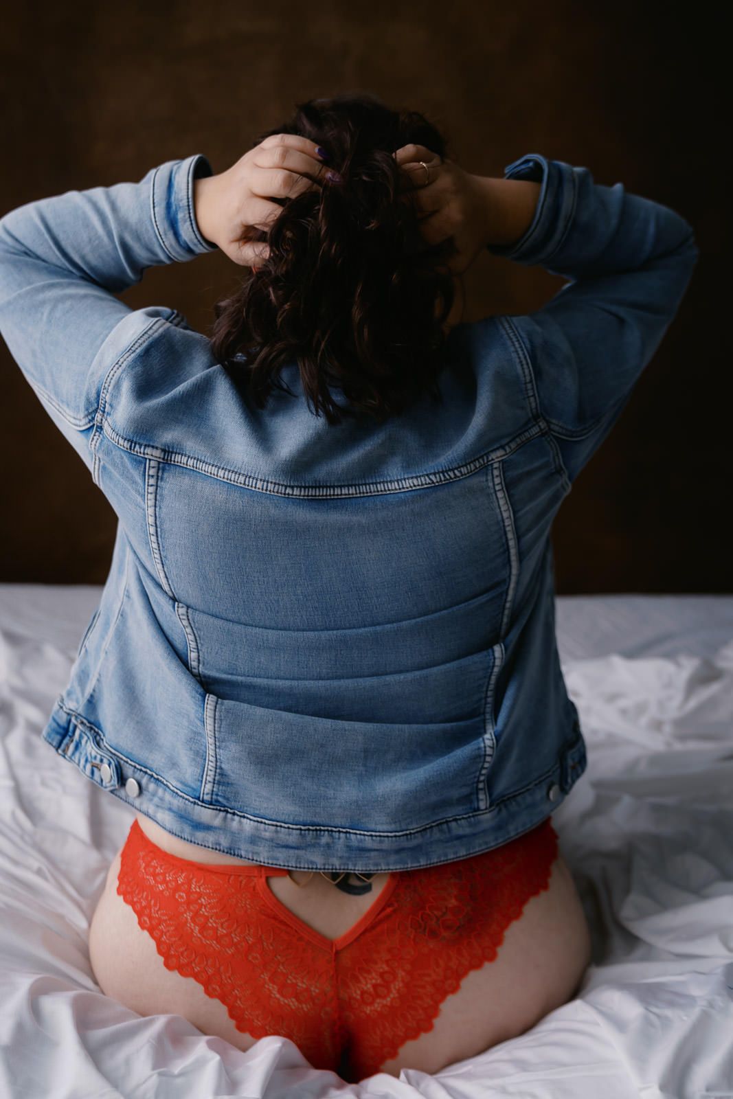 Woman in denim jacket and red lace underwear sitting on white bed, viewed from behind.