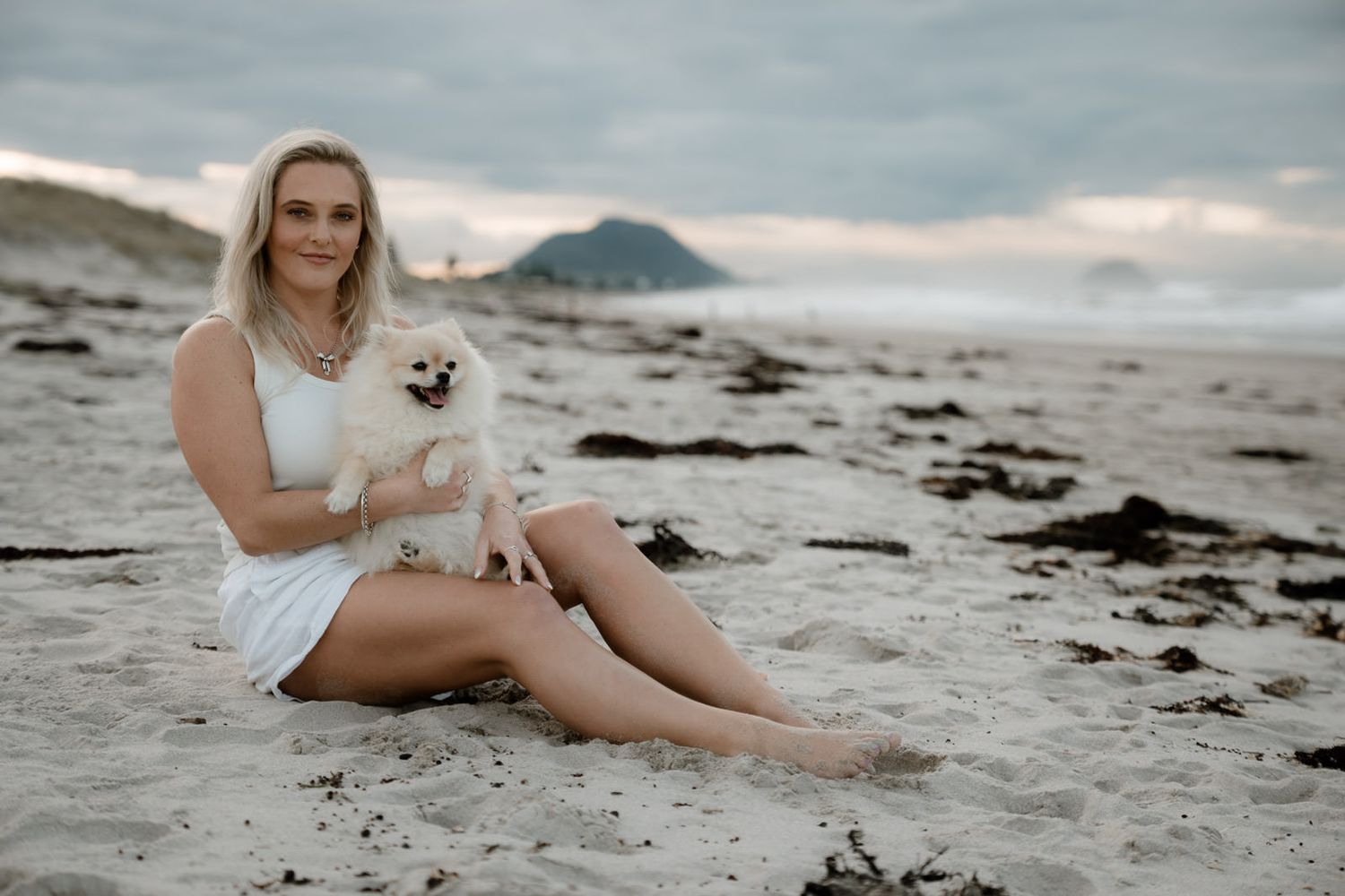 Woman in white dress sitting on beach holding fluffy white Pomeranian dog with rocky coastline backdrop.