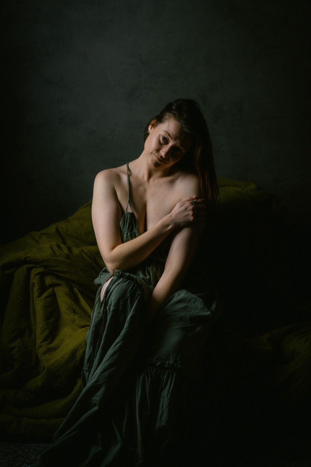 Woman in green silk dress sitting pensively against dark backdrop in moody fine art portrait photography.