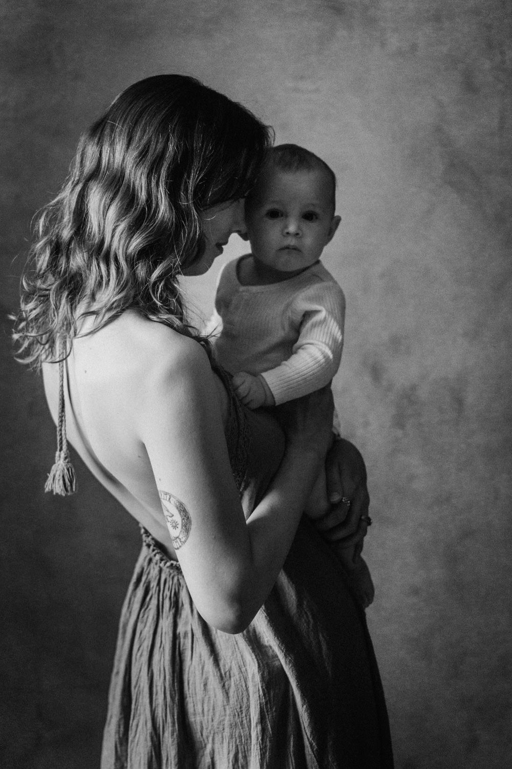 Black and white portrait of a mother holding her baby, with a tattoo visible on her arm, studio background.