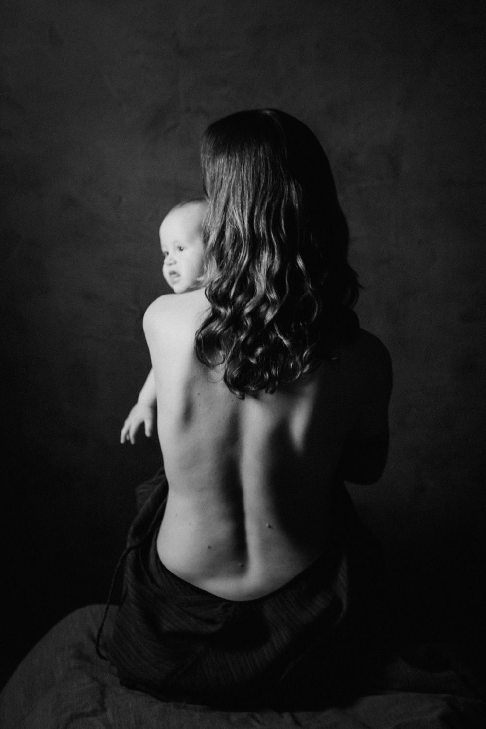 Black and white photo of a mother holding her baby, shot from behind, showing her bare back against a dark background.