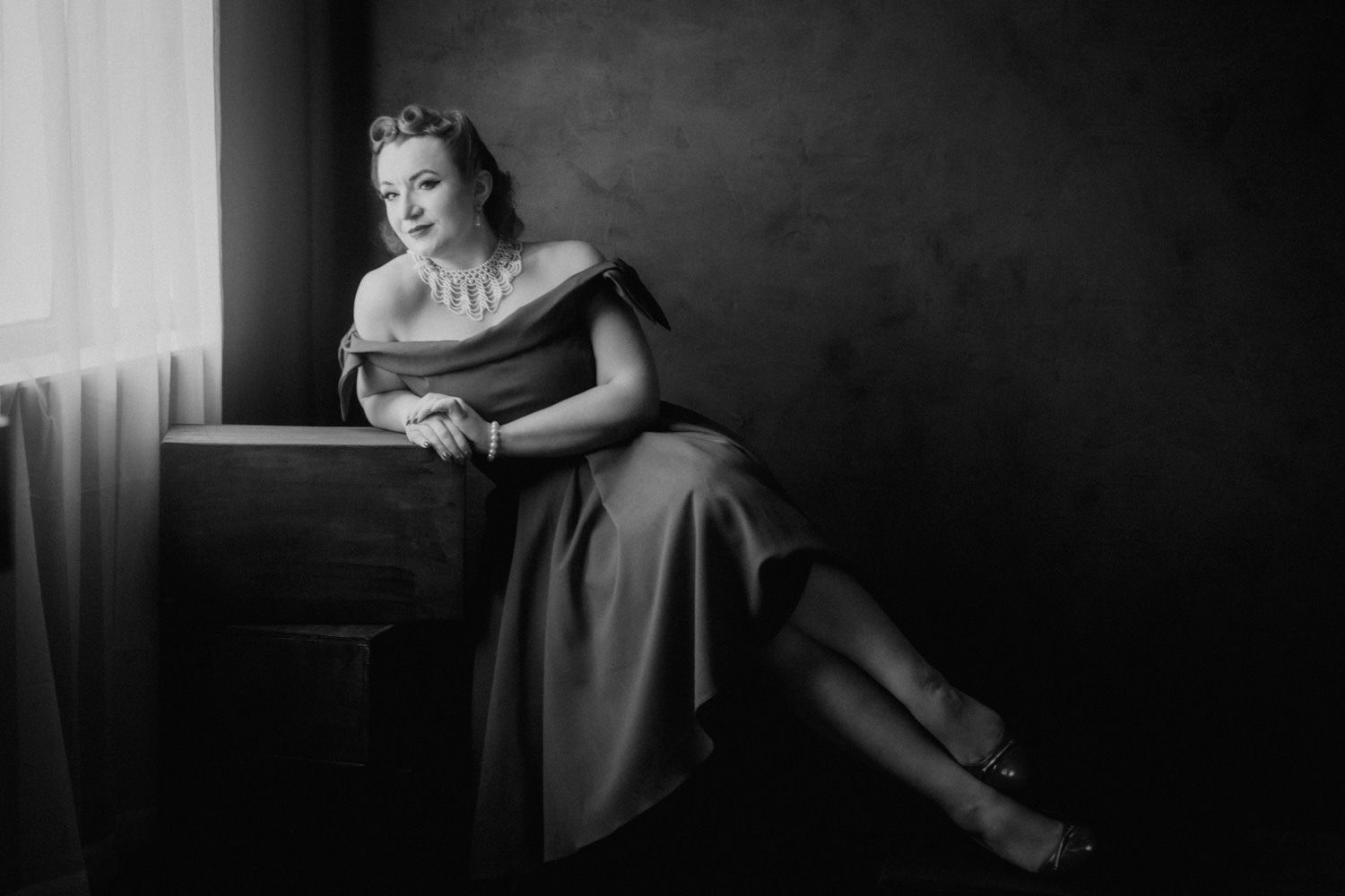 Elegant woman in off-shoulder gown poses gracefully by window in classic black and white portrait photography.