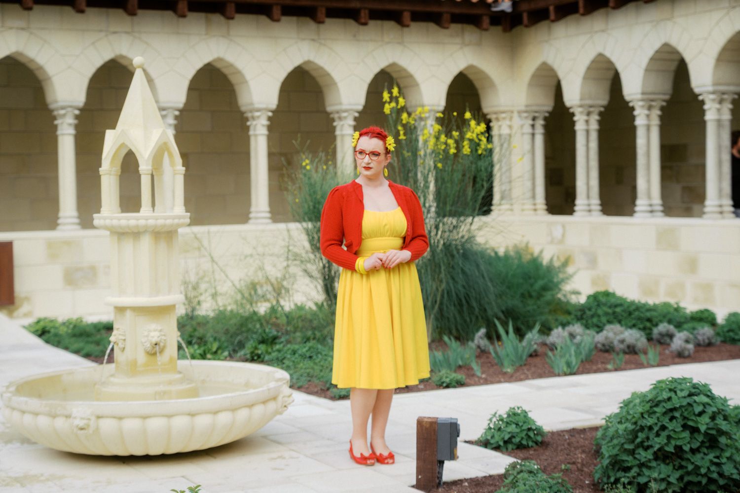 Woman in yellow dress and red cardigan stands by stone fountain in elegant courtyard with arched columns.